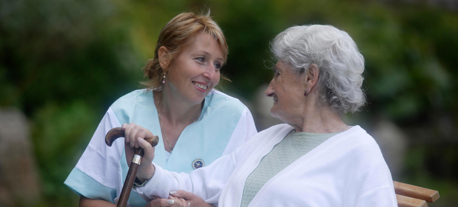 Alte Frau mit Gehstock mit Pflegerin auf einer Bank