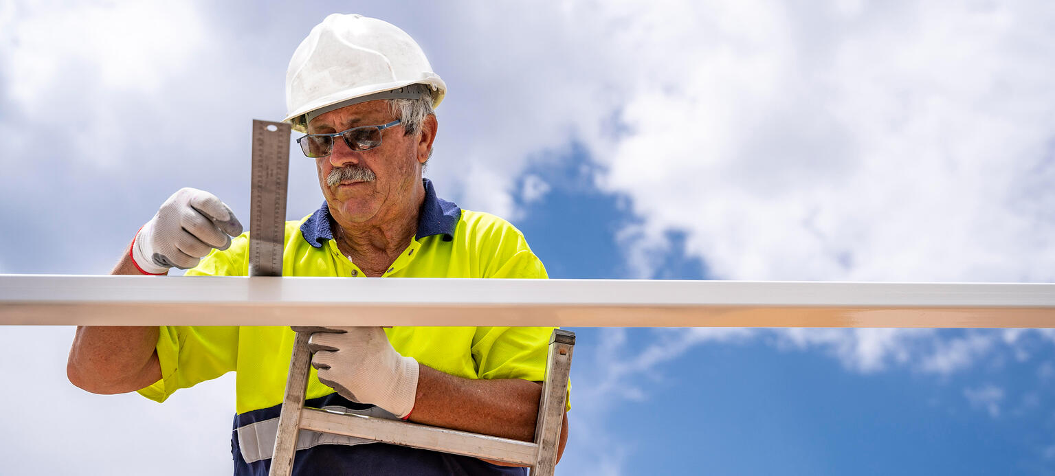 Senior male technician measuring roof while standing on ladder against cloudy sky 