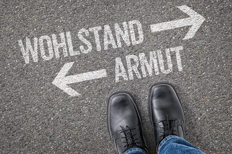Auf einer Straße stehen die Worte Wohlstand und Armut in weiß. Dort sind zwei weiße Pfeile, die in verschiedene Richtungen zeigen.