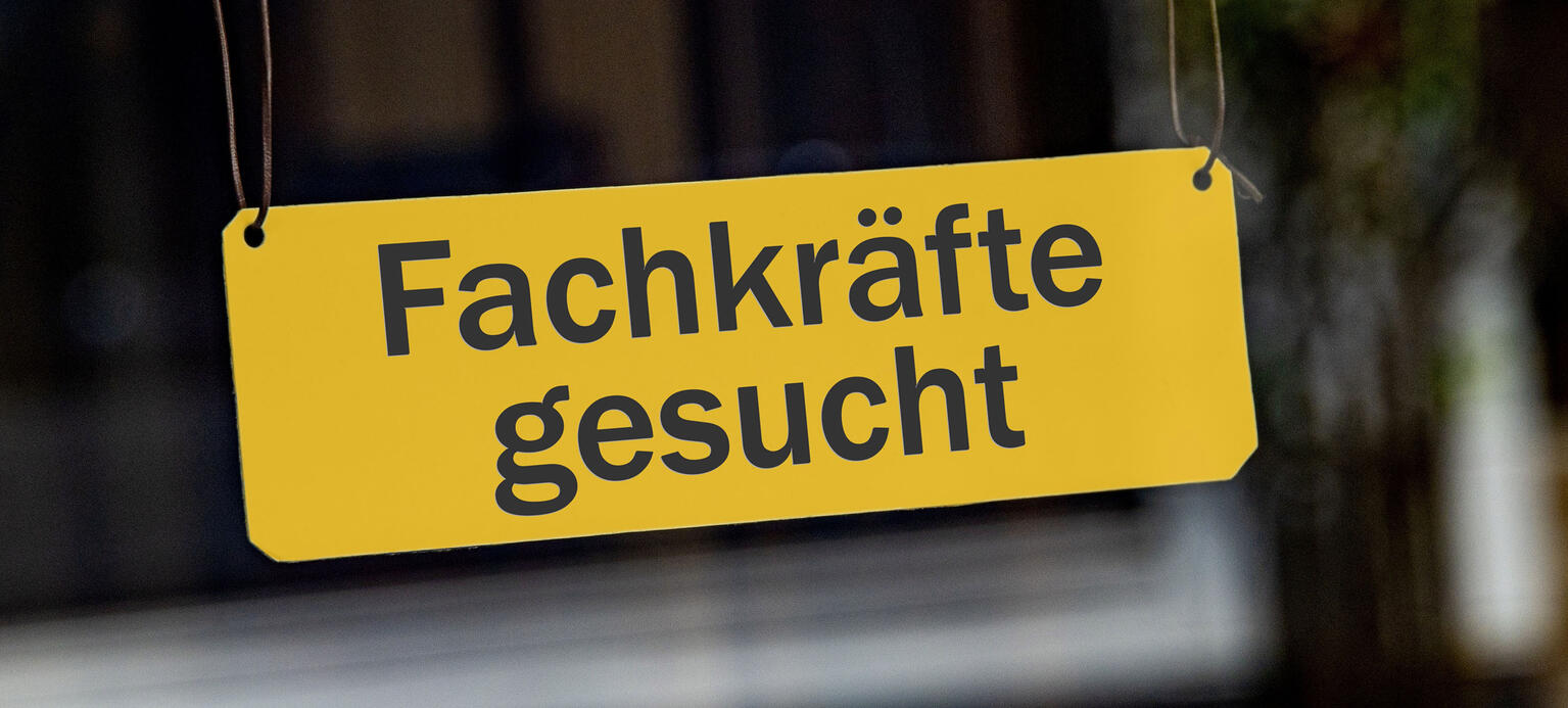 Ein symbolisches, gelbes Schild mit der Aufschrift 'Fachkraefte gesucht' haengt leicht schraeg an Lederbaendern in einem Schaufenster. 