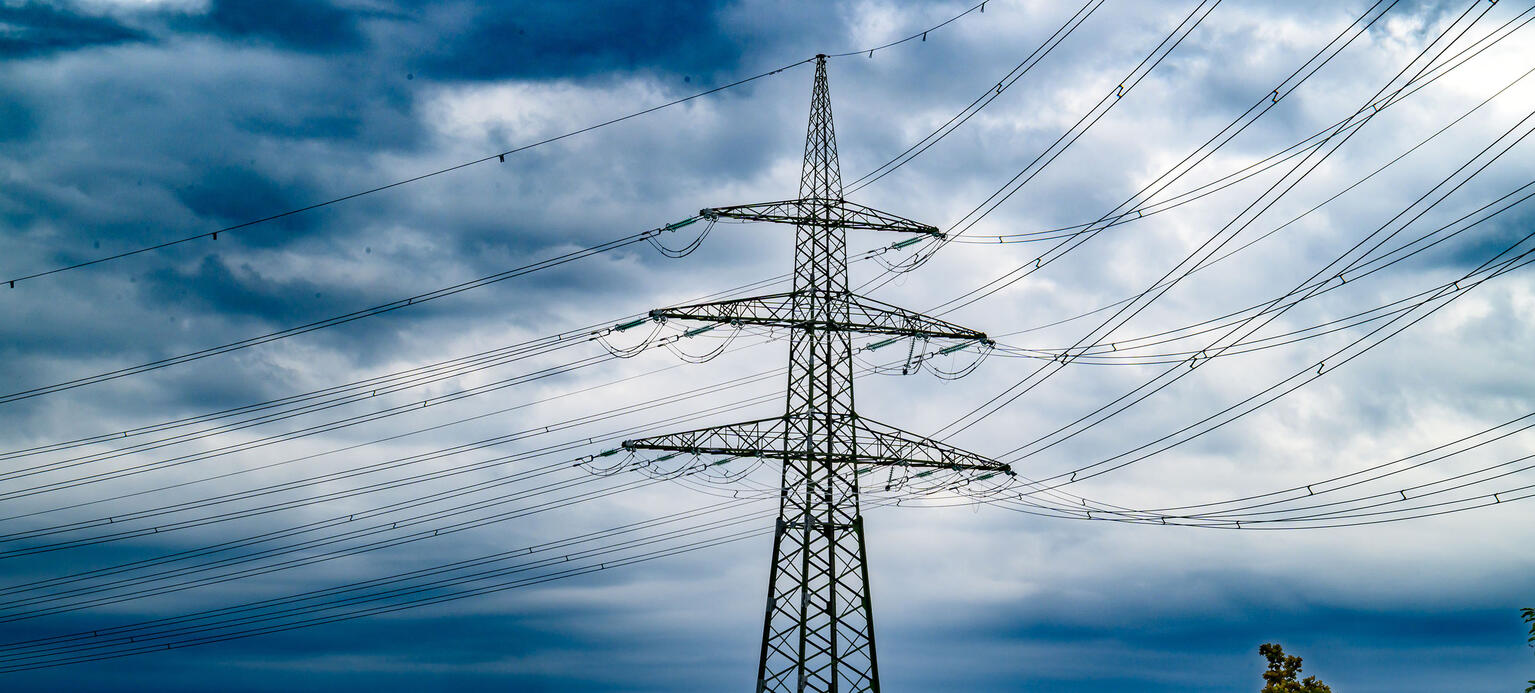 Eine Perspektive auf einen Hochspannungsmasten, der sich imposant vor einem teils dramatisch bewölkten Himmel abzeichnet. 