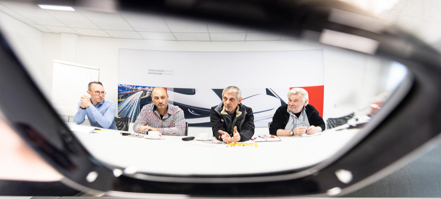 Blick in einen Seitenspiegel eines Autos, der von der Firma Magna gefertigt wurde. Darin spielt sich Christian Salomon, Georg Neuberger, Percy Scheidler und Heinz Pleßmann (v.l.n.r.) im Interview mit der Hans-Böckler-Stiftung 