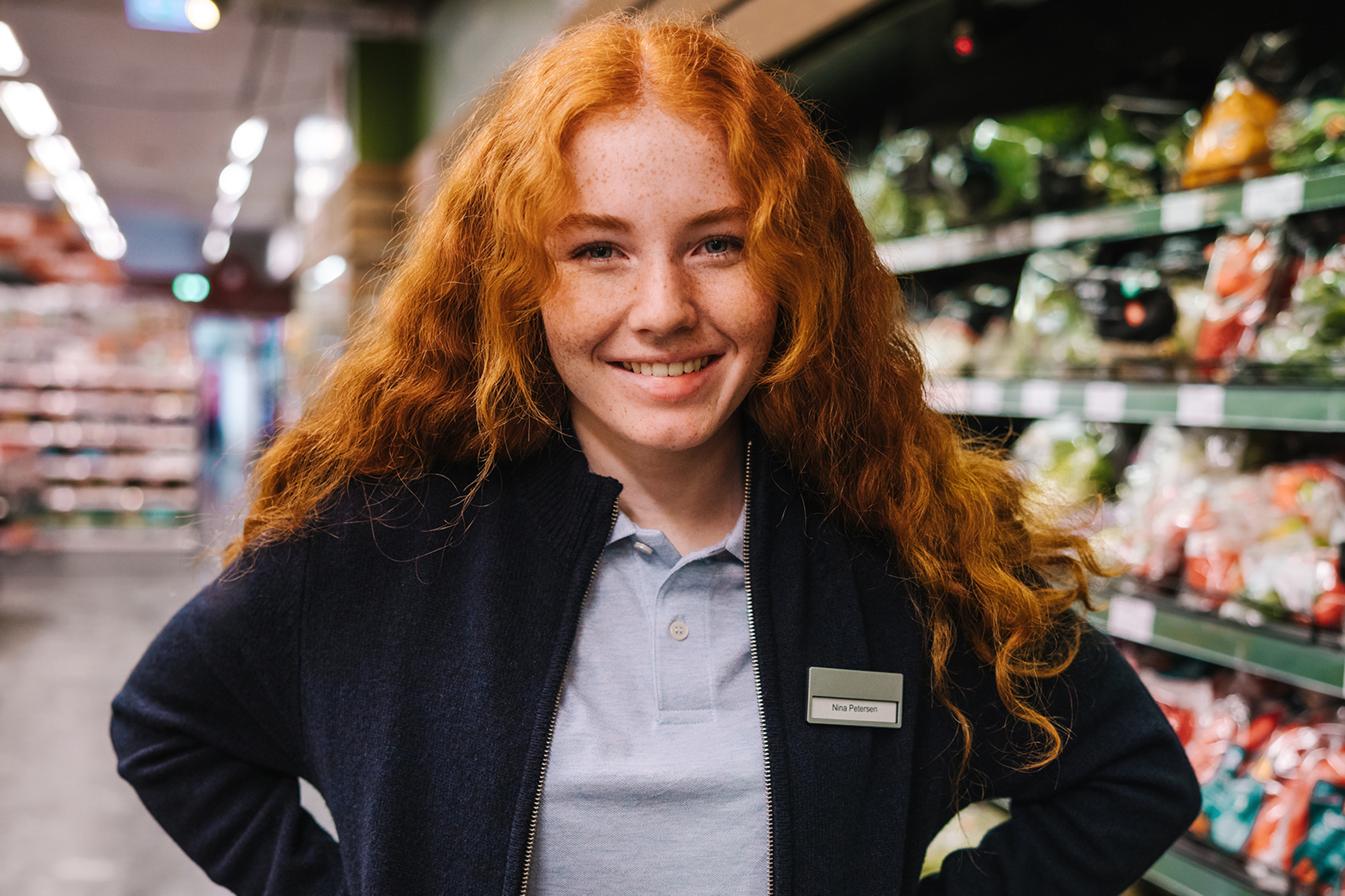 Lächelnde junge Frau (Azubi) steht im Supermarkt vor Obstregal